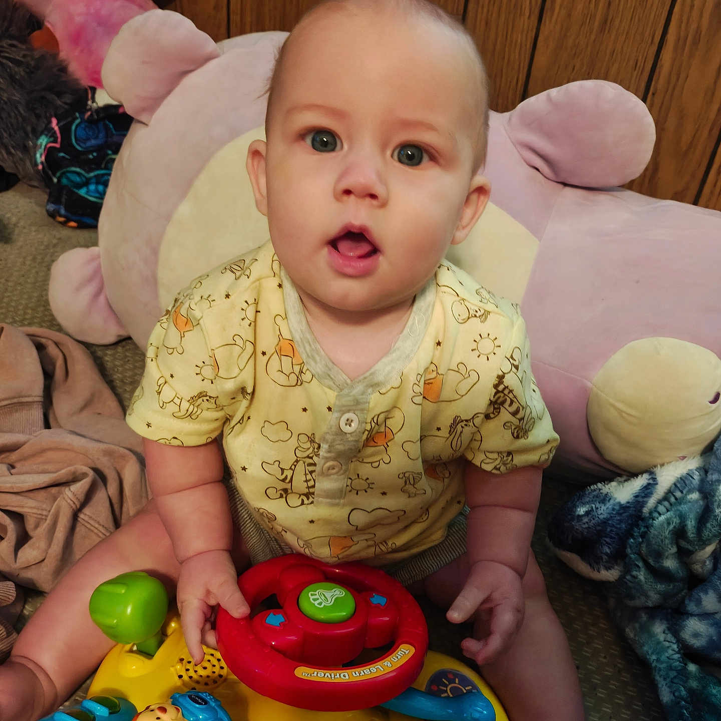 Wiliam is registered to the contest to win money with this photo: baby, carpet, child, colorful, cushion, cute, eyes, face, furniture, hands, indoor, infant, mouth_open, playtime, plush, sitting, steering_wheel, toy, wood_paneling, yellow_clothing