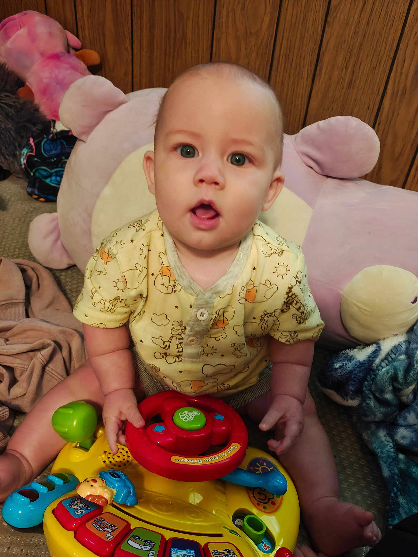 Wiliam is registered to the contest to win money with this photo: baby, child, toy, steering_wheel, plush, cushion, carpet, wood_paneling, yellow_clothing, sitting, indoor, hands, face, eyes, mouth_open, furniture, colorful, playtime, cute, infant