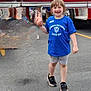 asphalt, blue_tshirt, casual_clothing, child, community_event, daytime, emergency_vehicle, fire_truck, happy, missing_tooth, outdoor, people, person, public_safety, rescue, road, shorts, smiling, standing, vehicle