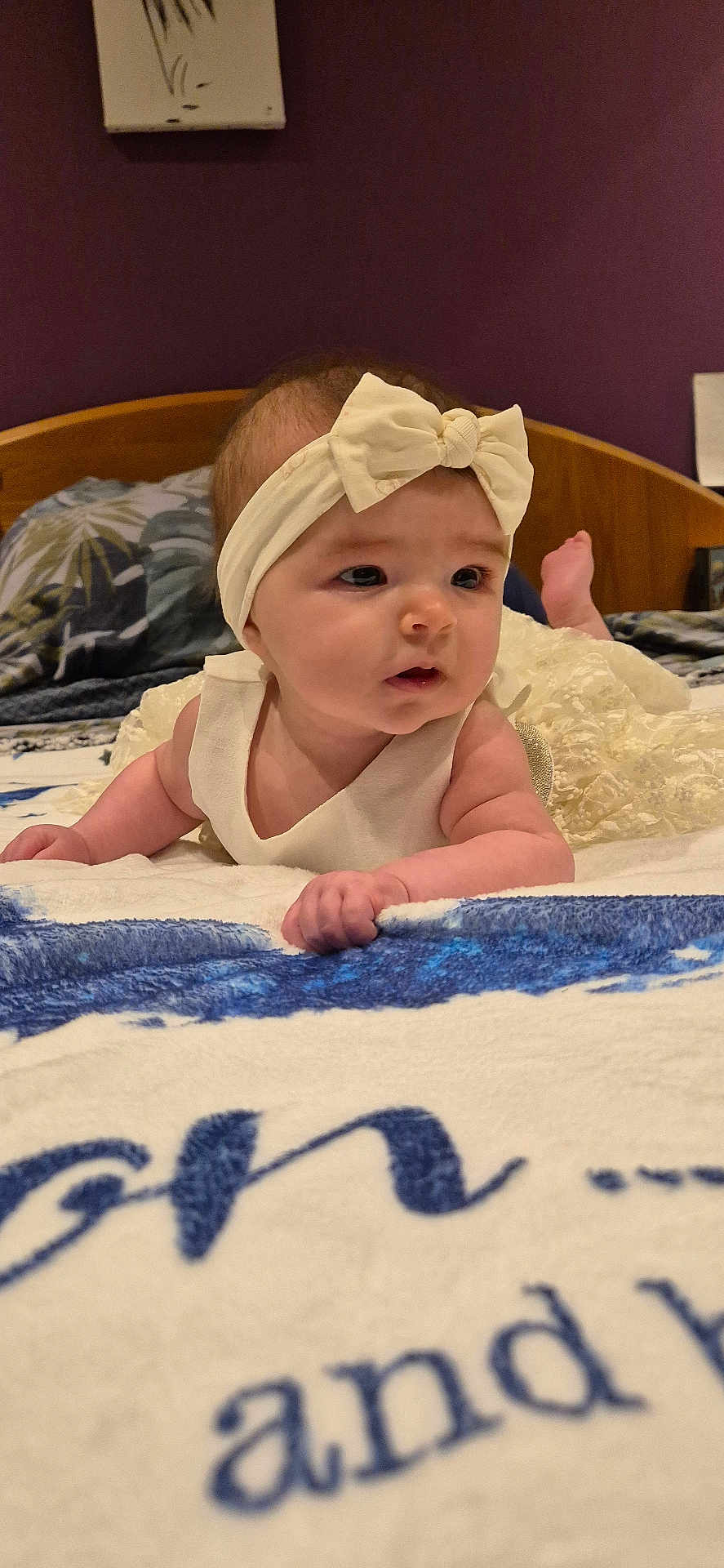 Aria a rejoint le concours — aidez-le/la à gagner de superbes lots ! baby, bed, bedding, blanket, bow, cheeks, cute, dress, eyes, face, hands, headband, indoor, infant, lying_on_stomach, pillow, portrait, purple_wall, toes, wooden_headboard