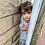 boy, casual_clothing, child, concrete_ground, curious, daylight, expression, hands, outdoor, peeking, person, playful, portrait, short_hair, side_view, sneakers, stone_wall, tiger_print_shirt, wooden_pole, young