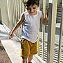 balancing, boy, casual_clothing, child, daylight, fence, happy, nature, outdoor, person, sandals, shadow, shorts, sidewalk, smile, summer, sunlight, walking, white_tank_top, young