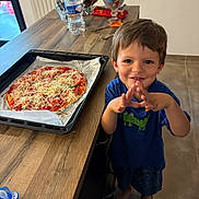 Jules a rejoint le concours — aidez-le/la à gagner de superbes lots ! child, boy, smiling, kitchen, pizza, baking_tray, countertop, water_bottle, candy, utensils, step_stool, barefoot, wooden_table, tile_floor, shorts, tshirt, food, homemade, indoor, happy