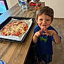 child, boy, smiling, kitchen, pizza, baking_tray, countertop, water_bottle, candy, utensils, step_stool, barefoot, wooden_table, tile_floor, shorts, tshirt, food, homemade, indoor, happy