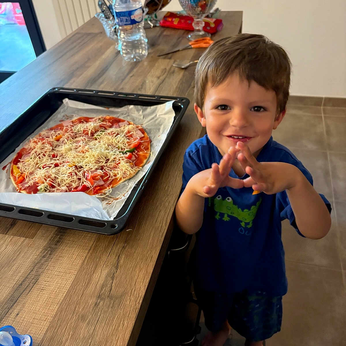 Jules a rejoint le concours — aidez-le/la à gagner de superbes lots ! baking_tray, barefoot, boy, candy, child, countertop, food, happy, homemade, indoor, kitchen, pizza, shorts, smiling, step_stool, tile_floor, tshirt, utensils, water_bottle, wooden_table