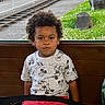 child, toddler, curly_hair, mickey_mouse, shirt, red_pants, bench, seat, green_chair, window, outdoor, railway_track, grass, plants, fence, car, serious_expression, portrait, indoors, casual_clothing