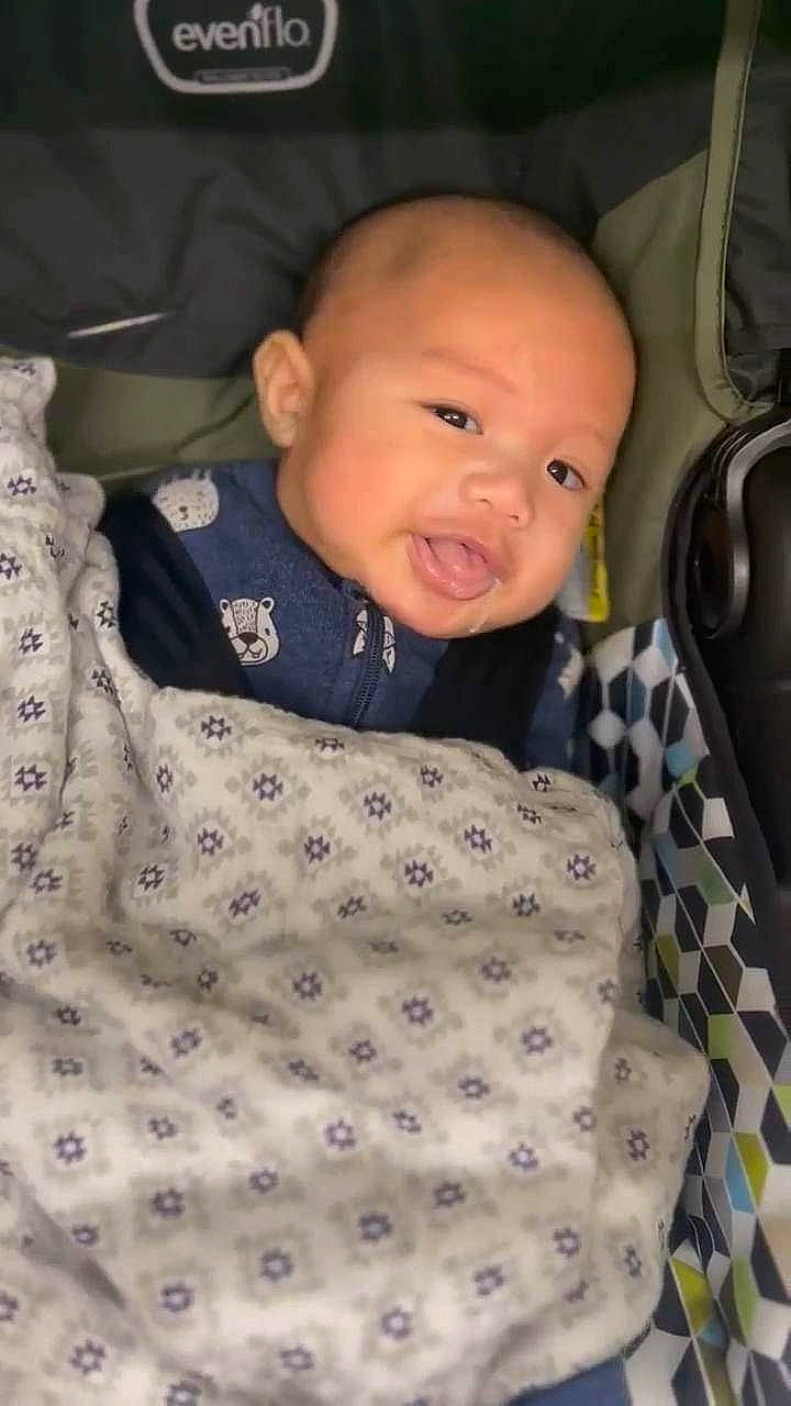 Kian is registered to the contest to win money with this photo: baby, baby_products, baby_safety, baby_toddler_clothing, car_seat, cheek, child, collar, comfort, linens, pattern, person, room, sitting, sleeve, smile, textile, toddler