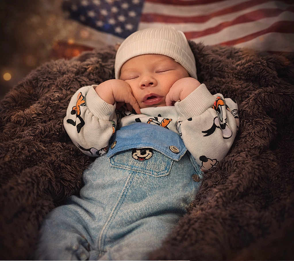 Maël a rejoint le concours — aidez-le/la à gagner de superbes lots ! baby, sleeping, infant, denim_overalls, white_hat, blanket, brown_fur, american_flag, cozy, cute, peaceful, portrait, child, soft_light, indoors, clothing, hands, face, resting, newborn