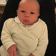 Maël a rejoint le concours — aidez-le/la à gagner de superbes lots ! baby, infant, white_clothing, sitting, lap, hands, face, eyes, tongue, indoor, wooden_table, person, knitwear, soft_lighting, cozy, casual, portrait, young_child, closeup, home