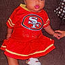 baby, child, infant, portrait, sitting, couch, red_outfit, football_jersey, sportswear, san_francisco_49ers, headband, bow, sandals, bracelet, smiling, wide_eyed, indoor, furniture, fashion, football_fan
