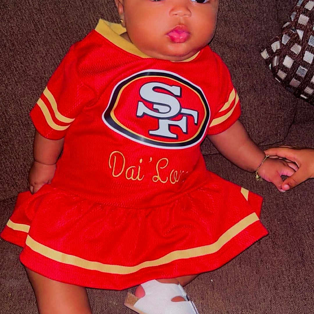 Dlonni is registered to the contest to win money with this photo: baby, child, infant, portrait, sitting, couch, red_outfit, football_jersey, sportswear, san_francisco_49ers, headband, bow, sandals, bracelet, smiling, wide_eyed, indoor, furniture, fashion, football_fan