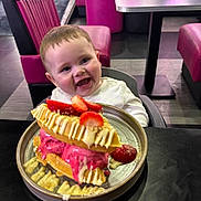 George is registered to the contest to win money with this photo: baby, child, high_chair, waffle, ice_cream, strawberry, dessert, plate, table, diner, pink_chair, smiling, happy, food, indoor, person, sweet, cream, drizzle, seat