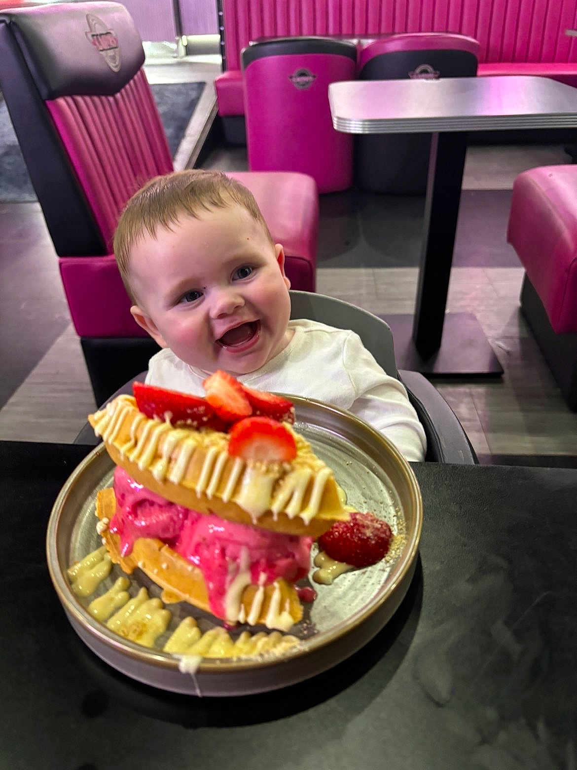 George is registered to the contest to win money with this photo: baby, child, high_chair, waffle, ice_cream, strawberry, dessert, plate, table, diner, pink_chair, smiling, happy, food, indoor, person, sweet, cream, drizzle, seat