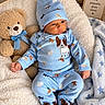 baby, infant, child, portrait, indoors, blanket, bed, teddy_bear, stuffed_animal, blue_clothing, pajamas, baby_hat, baby_shoes, dog_pattern, cute, sitting, lying_down, nursery, cozy, framed_sign