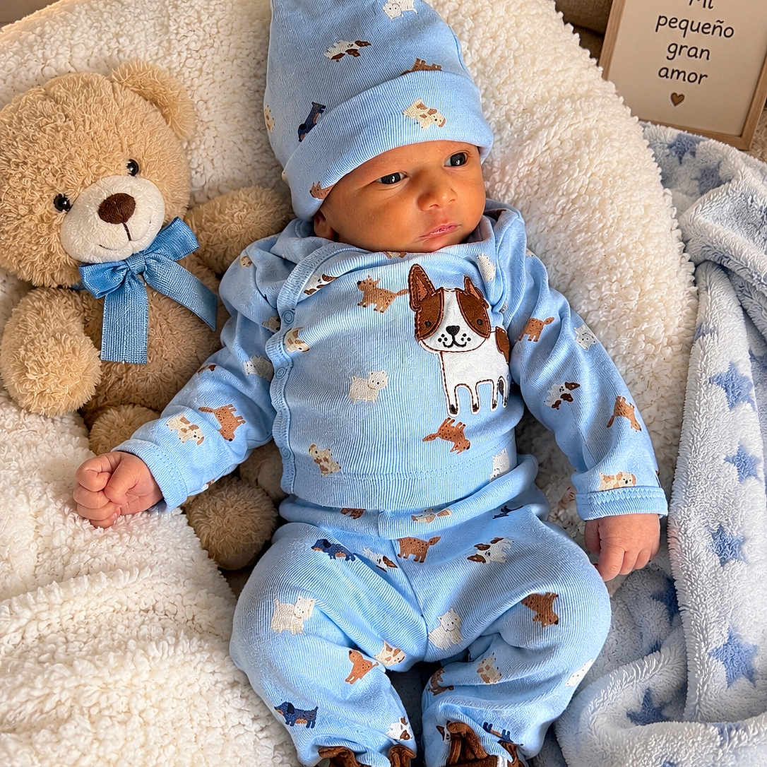 Chris Marco is registered to the contest to win money with this photo: baby, infant, child, portrait, indoors, blanket, bed, teddy_bear, stuffed_animal, blue_clothing, pajamas, baby_hat, baby_shoes, dog_pattern, cute, sitting, lying_down, nursery, cozy, framed_sign