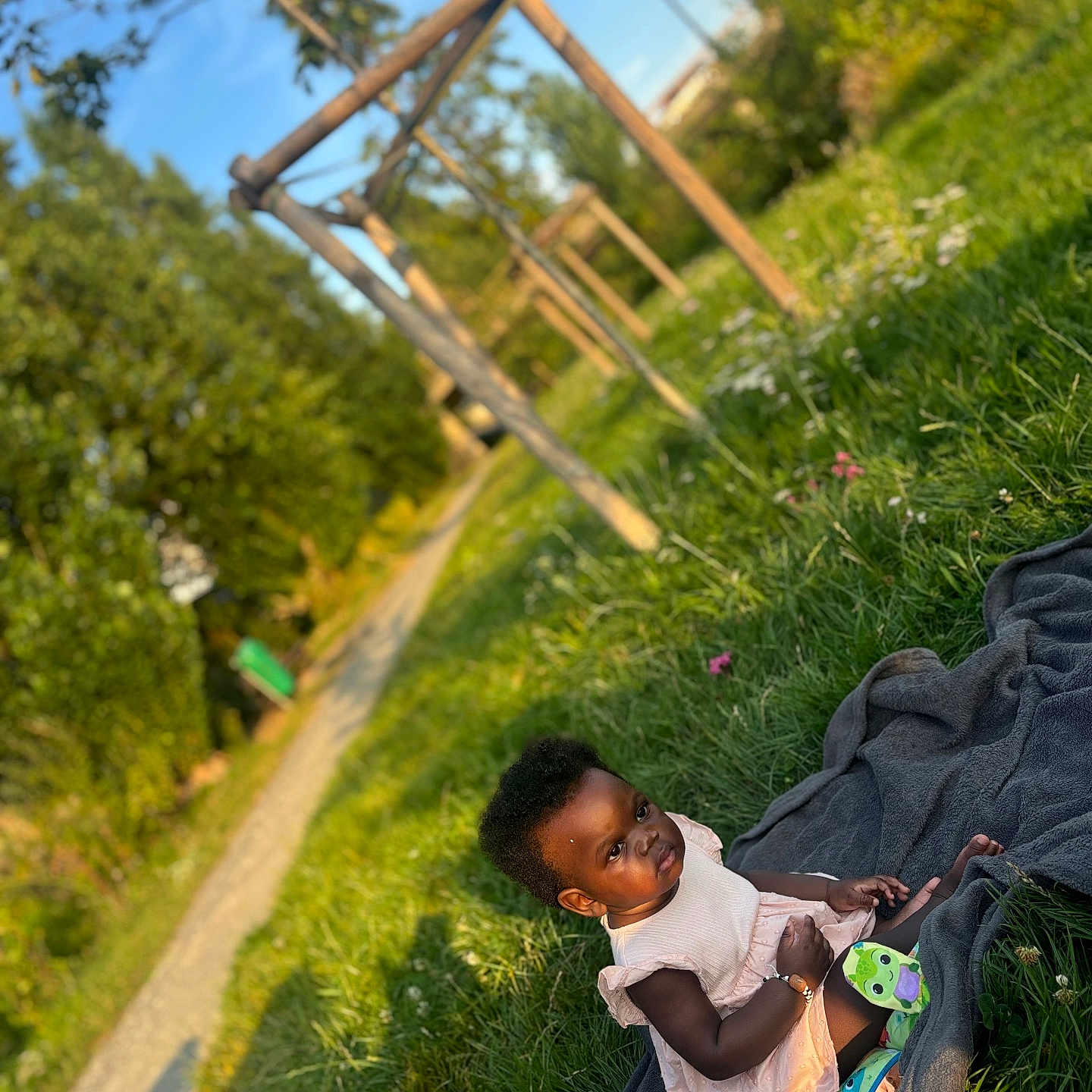 Mencia a rejoint le concours — aidez-le/la à gagner de superbes lots ! blanket, child, cute, daylight, dress, grass, greenery, nature, outdoor, park, pathway, person, playground, sitting, socks, summer, sunlight, toddler, wooden_structure, young_child