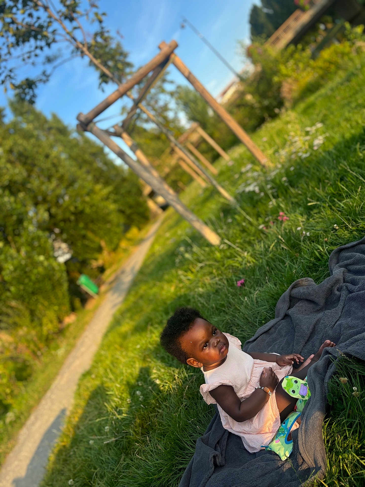 Mencia a rejoint le concours — aidez-le/la à gagner de superbes lots ! child, toddler, grass, outdoor, blanket, dress, socks, pathway, wooden_structure, sunlight, greenery, nature, playground, person, summer, daylight, park, cute, young_child, sitting