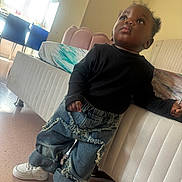 Mencia participe au concours pour gagner de l'argent avec cette photo : bed, black_shirt, casual_clothing, child, curious_expression, curly_hair, daylight, denim, floor, furniture, home, indoor, jeans, leaning, person, standing, toddler, white_sneakers, window, young_child