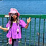 child, girl, hat, pink_hat, sunglasses, purple_top, pants, standing, fence, lake, water, outdoor, sunny, daylight, playful, summer, accessory, fashion, person, casual