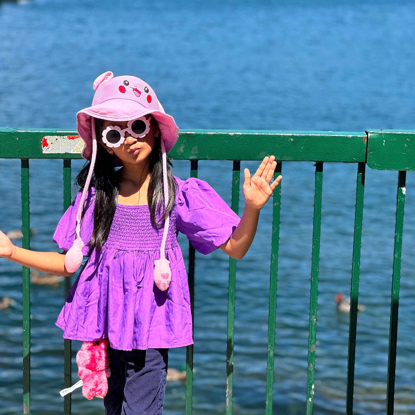 Rushda is registered to the contest to win money with this photo: accessory, casual, child, daylight, fashion, fence, girl, hat, lake, outdoor, pants, person, pink_hat, playful, purple_top, standing, summer, sunglasses, sunny, water