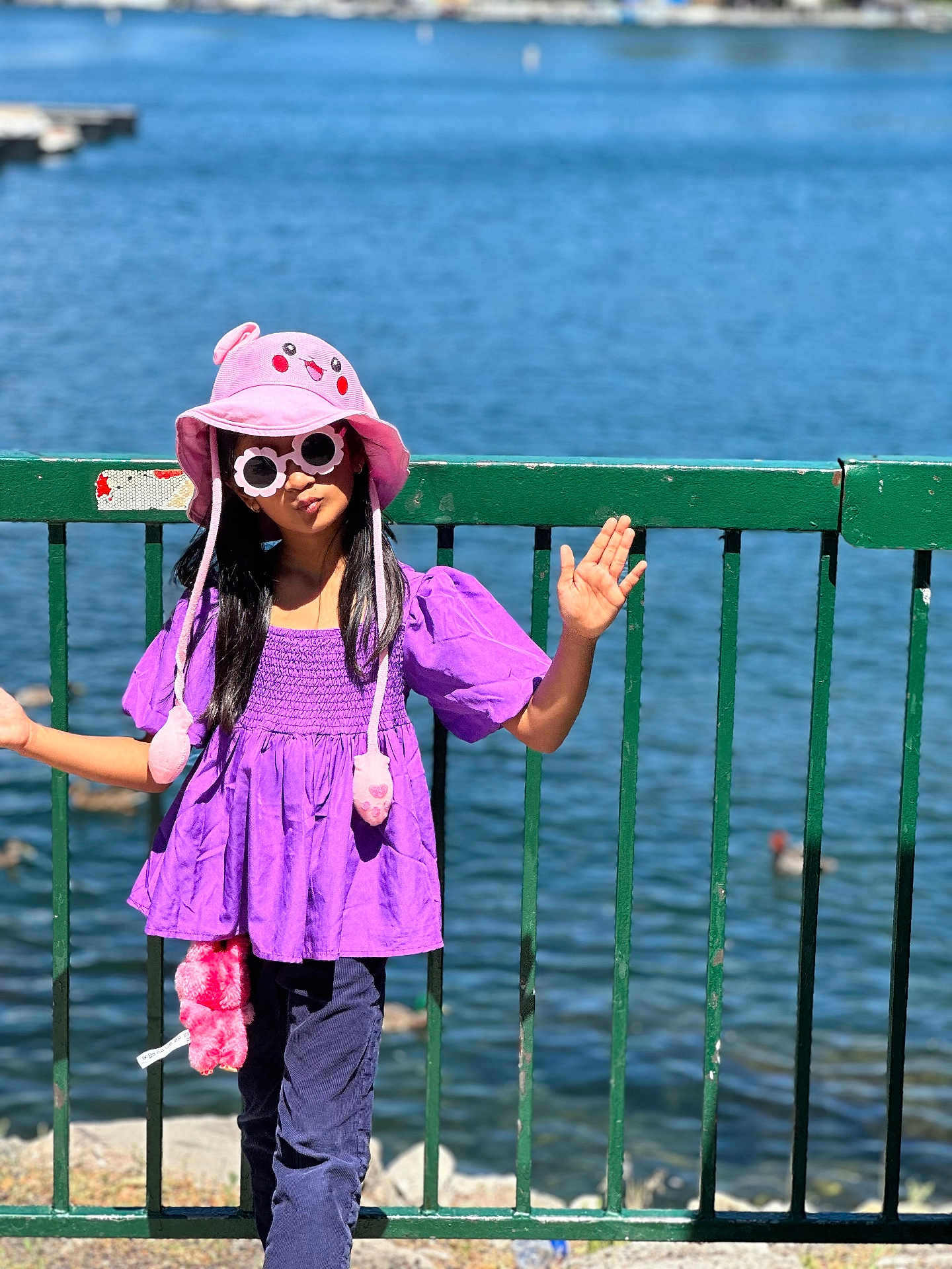 Rushda is registered to the contest to win money with this photo: child, girl, hat, pink_hat, sunglasses, purple_top, pants, standing, fence, lake, water, outdoor, sunny, daylight, playful, summer, accessory, fashion, person, casual