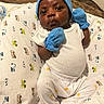 baby, infant, blue_mittens, blue_hat, white_onesie, white_pants, blanket, patterned_blanket, cute, portrait, indoors, child, newborn, clothing, sleepwear, pouty_face, small_hands, sitting, soft_lighting, cozy
