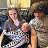 baby, child, teenager, couch, leather, siblings, smile, earbuds, long_hair, curly_hair, plaid_pajamas, indoor, window, air_conditioner, casual_clothing, family, relaxed, portrait, seated, wood_paneling