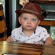 Raymond joined the competition — help win amazing prizes! blue_eyes, brown_hat, casual_clothing, child, closeup, curious_expression, cute, face, furniture, hat, indoor, menu, person, portrait, restaurant, sitting, table, toddler, white_polo_shirt, wooden_table