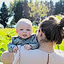 baby, child, person, adult, hair_bun, glasses, earrings, checkered_shirt, outdoor, nature, greenery, trees, sunlight, shoulder, holding, tongue_out, face, head, daylight, field