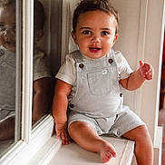 Chandler is registered to the contest to win money with this photo: child, toddler, smiling, indoor, reflection, window, overalls, white_shirt, curly_hair, happy, face, sitting, barefoot, skin, light, window_ledge, portrait, young_child, cute, person
