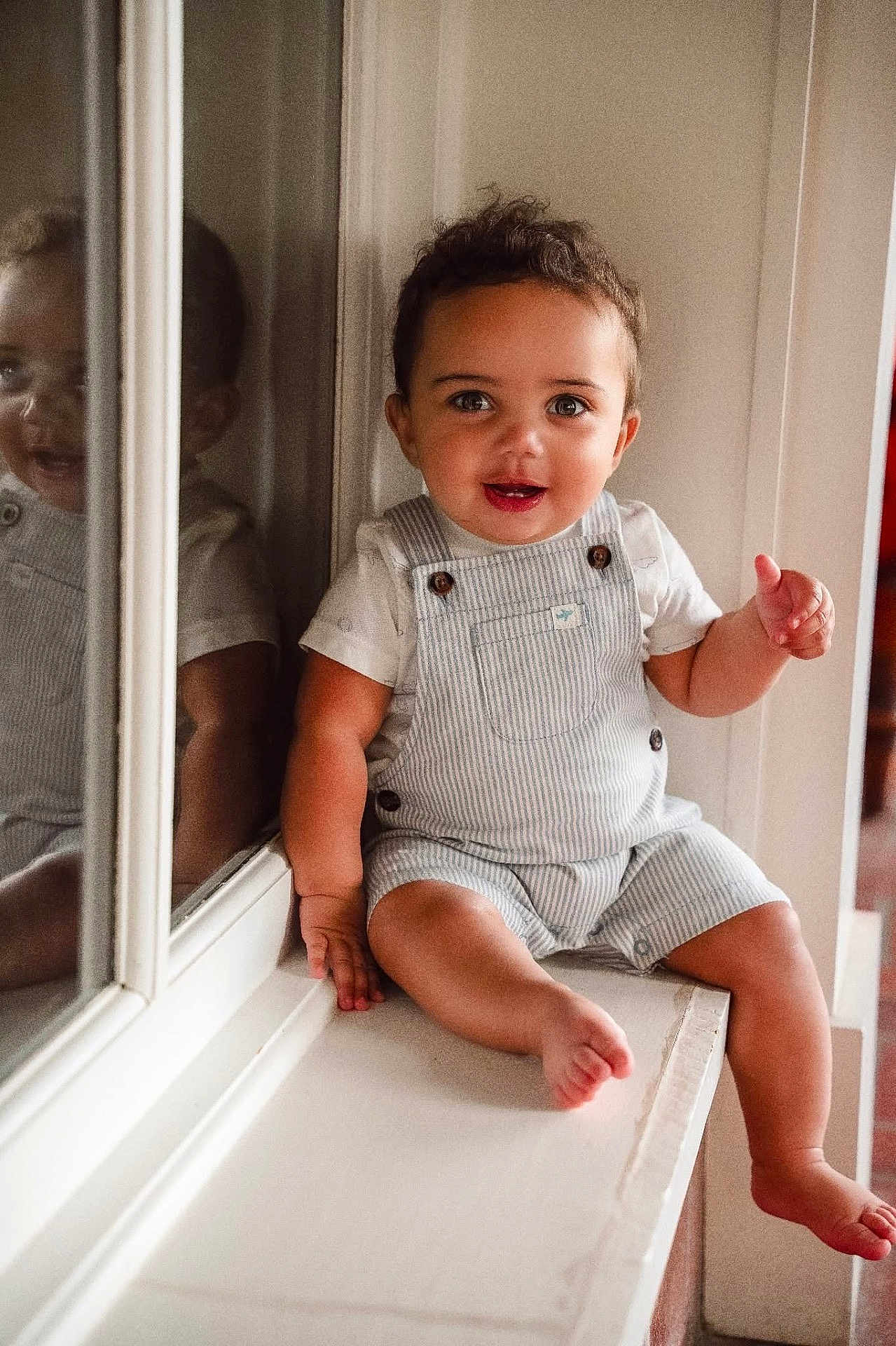 Chandler is registered to the contest to win money with this photo: child, toddler, smiling, indoor, reflection, window, overalls, white_shirt, curly_hair, happy, face, sitting, barefoot, skin, light, window_ledge, portrait, young_child, cute, person