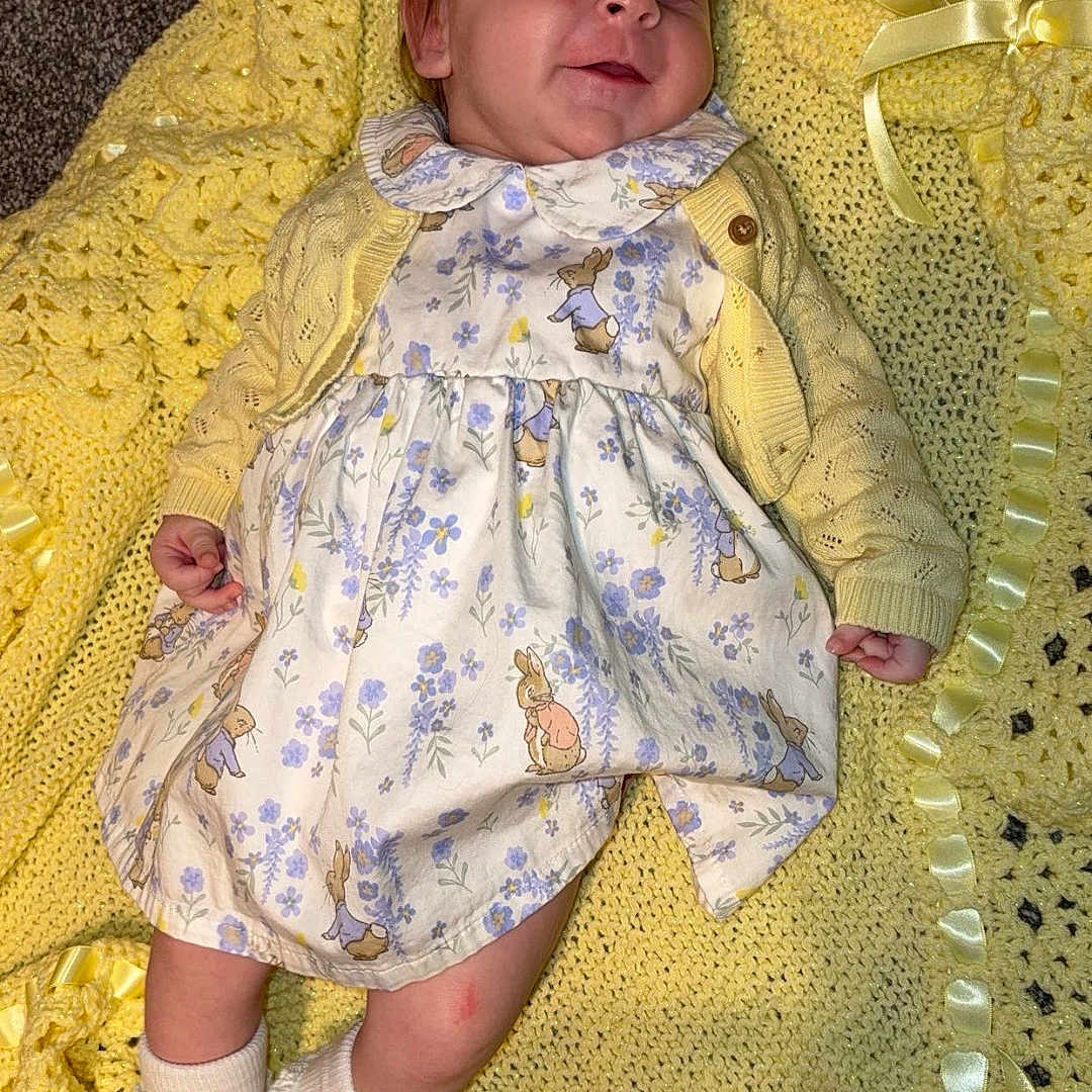 Dolores is registered to the contest to win money with this photo: baby, cardigan, carpet, child, comfortable, crochet, cute, dress, floor, floral_pattern, happy, indoors, infant, lying_down, portrait, ribbons, smiling, socks, soft_texture, yellow_blanket