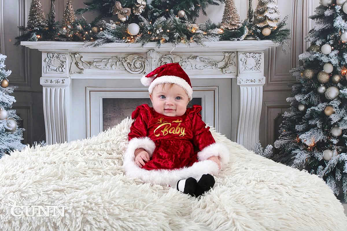 Lily is registered to the contest to win money with this photo: baby, christmas, holiday, festive, santa_hat, red_dress, fireplace, decorations, christmas_tree, ornaments, blanket, smiling, cute, indoor, winter, celebration, child, portrait, cozy, festivity