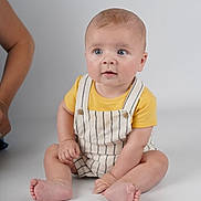 Thomas joined the competition — help win amazing prizes! baby, child, sitting, yellow_shirt, striped_overalls, blue_eyes, curious, indoor, studio, white_background, barefoot, person, infant, cute, portrait, skin, hand, foot, head, casual_clothing
