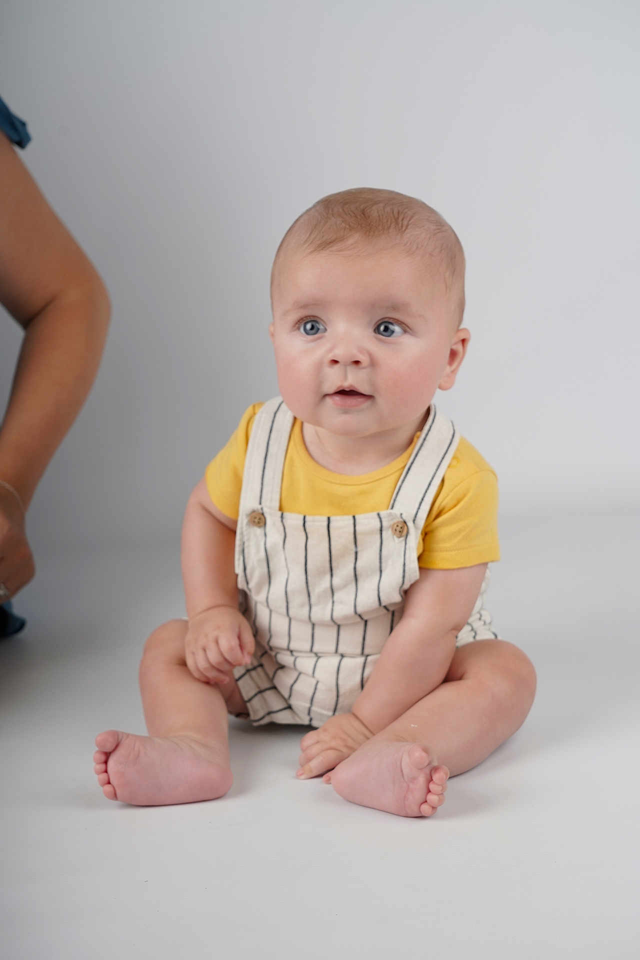 Thomas joined the competition — help win amazing prizes! baby, child, sitting, yellow_shirt, striped_overalls, blue_eyes, curious, indoor, studio, white_background, barefoot, person, infant, cute, portrait, skin, hand, foot, head, casual_clothing