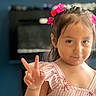 child, girl, portrait, smiling, peace_sign, flower_clips, pink_dress, striped_clothing, long_hair, indoors, close_up, face, eyes, hand_gesture, happy, looking_at_camera, blurred_background, casual, photography, youth