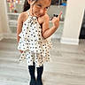 child, girl, toddler, portrait, indoors, smiling, side_eye, thumbs_up, polka_dot_dress, dress, black_tights, ponytail, fashion, standing, wood_floor, home, shelves, furniture, playful, photography