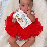 Delilah joined the competition — help win amazing prizes! baby, child, tutu, red_clothing, white_blanket, card, giraffe, months_old, infant, cute, lying_down, hand_near_mouth, curious, wide_eyes, soft_texture, indoors, floor_rug, patterned_rug, colorful, portrait