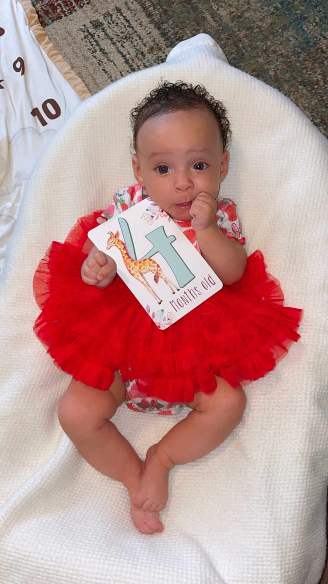 Delilah joined the competition — help win amazing prizes! baby, child, tutu, red_clothing, white_blanket, card, giraffe, months_old, infant, cute, lying_down, hand_near_mouth, curious, wide_eyes, soft_texture, indoors, floor_rug, patterned_rug, colorful, portrait