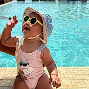 Elyne a rejoint le concours — aidez-le/la à gagner de superbes lots ! baby, child, sunglasses, hat, swimsuit, pool, water, sunny, vacation, smiling, happy, waving, outdoor, summer, tiled_edge, ocean, umbrella, people, background, sunlight