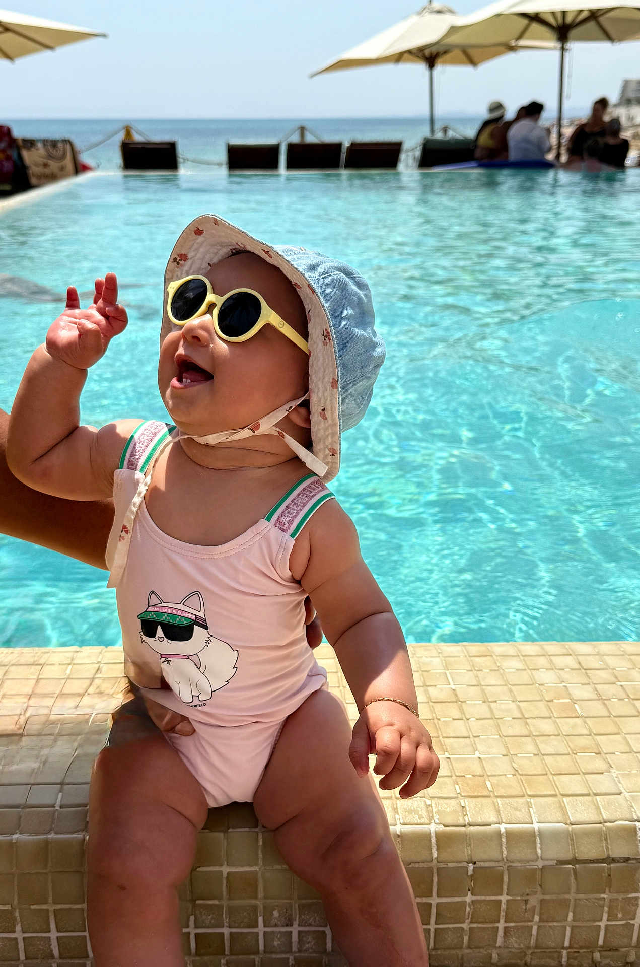 Elyne a rejoint le concours — aidez-le/la à gagner de superbes lots ! baby, child, sunglasses, hat, swimsuit, pool, water, sunny, vacation, smiling, happy, waving, outdoor, summer, tiled_edge, ocean, umbrella, people, background, sunlight