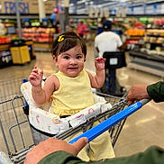 Luna is registered to the contest to win money with this photo: baby, child, shopping_cart, grocery_store, yellow_clothing, smiling, happy, indoor, person, shopping, seat_cover, rainbow_pattern, bracelet, hair_clip, retail, aisle, hand, mobility_scooter, background, store_shelves