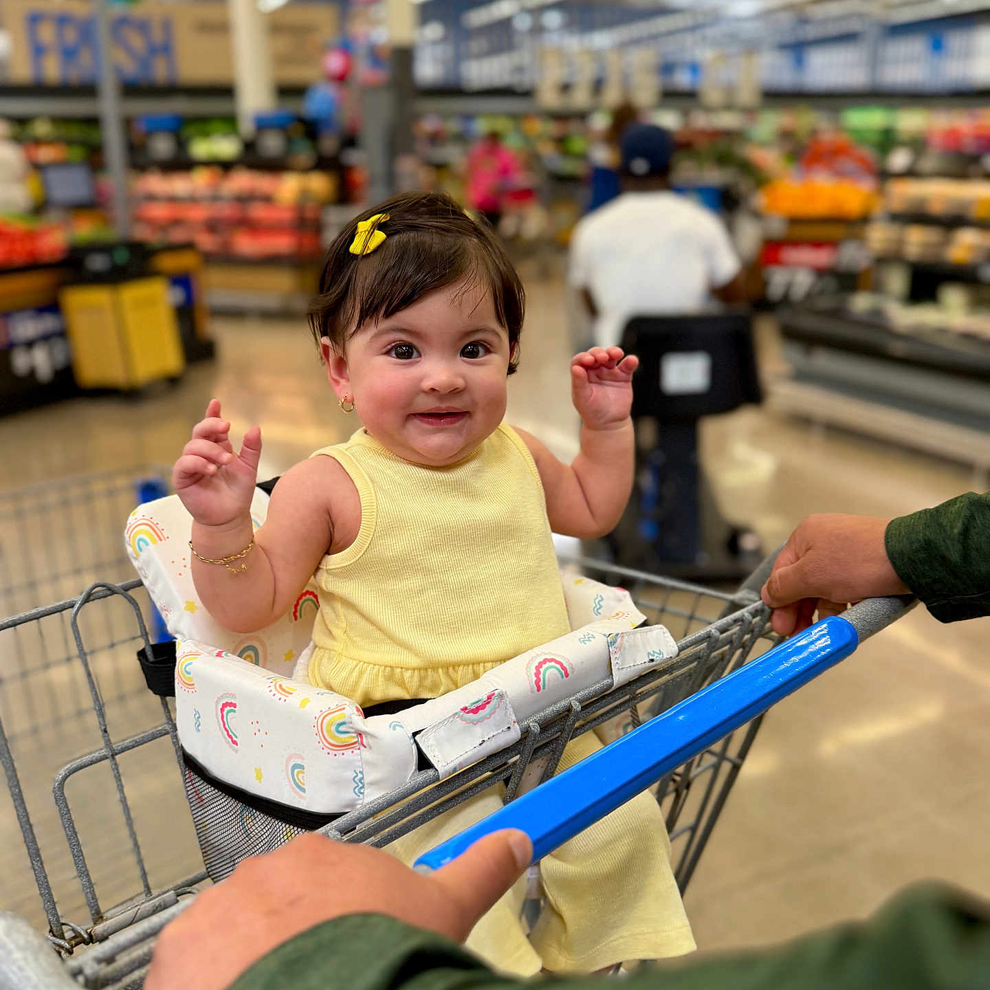 Luna is registered to the contest to win money with this photo: baby, child, shopping_cart, grocery_store, yellow_clothing, smiling, happy, indoor, person, shopping, seat_cover, rainbow_pattern, bracelet, hair_clip, retail, aisle, hand, mobility_scooter, background, store_shelves