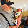 Alden is registered to the contest to win money with this photo: baby, infant, man, adult, beard, cap, hat, holding, cradle, tattoo, suspenders, tshirt, smile, home_interior, wall_outlet, chair, table, jeans, wood_floor, portrait
