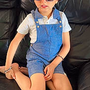 Manan is registered to the contest to win money with this photo: child, boy, sunglasses, blue_hat, blue_overalls, white_shirt, bracelet, black_couch, indoor, fashion, sitting, cross_legged, casual, portrait, cute, young, person, leather_furniture, relaxed, accessory