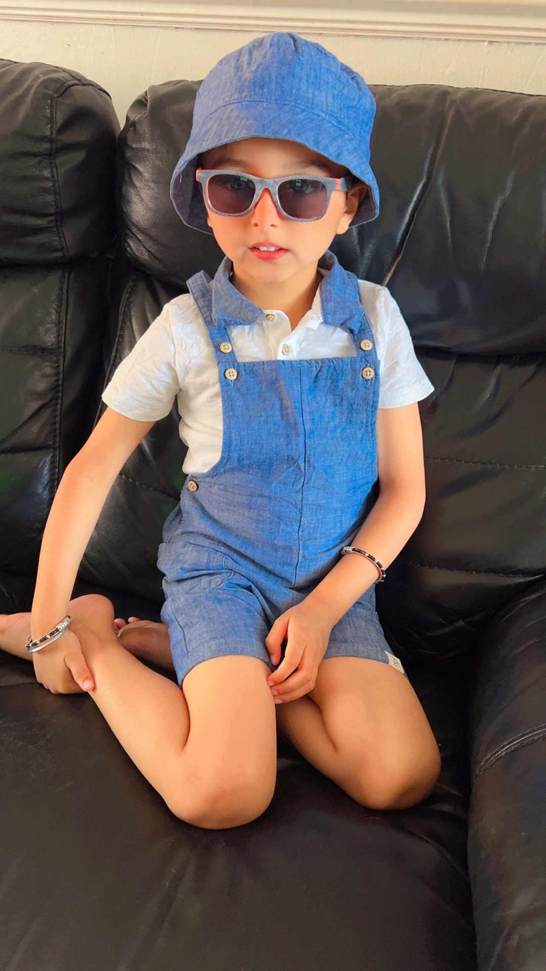 Manan is registered to the contest to win money with this photo: child, boy, sunglasses, blue_hat, blue_overalls, white_shirt, bracelet, black_couch, indoor, fashion, sitting, cross_legged, casual, portrait, cute, young, person, leather_furniture, relaxed, accessory