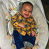 baby, child, smiling, yellow_shirt, plaid, jeans, brown_shoes, baby_seat, cushion, indoor, happy, person, cute, face, infant, portrait, clothing, footwear, seated, tongue_out