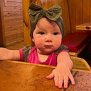 Carter joined the competition — help win amazing prizes! baby, bench, bow, candid, child, curious, cute, expression, face, hand, headband, indoor, person, pink_clothing, portrait, seated, table, wood_paneling, wooden_table, young_child