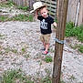 child, cowboy_hat, tshirt, shorts, shoes, wooden_post, chain, grass, dirt, fence, wooden_shed, outdoor, person, standing, rural, yard, summer, cute, playful, hat