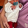 Ellie participe au concours pour gagner de l'argent avec cette photo : baby, newborn, pacifier, pink_hat, white_sweater, red_pants, blue_slippers, cartoon_character, sleeping, adult_arm, purple_shirt, nike_logo, hand, cozy_clothing, indoor, close_up, peaceful, cradling, infant, cute